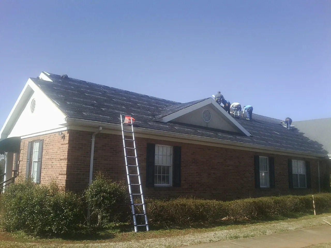 Crew tearing shingles off a brick ranch roof with a ladder set — Metal Roof Repair work by our crew in Moncure
