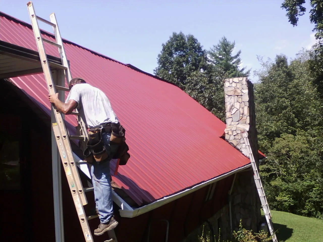 Roofer on a ladder beside a red metal roof and stone chimney — Soffit & Fascia Repair by Roofing Brothers in Moncure